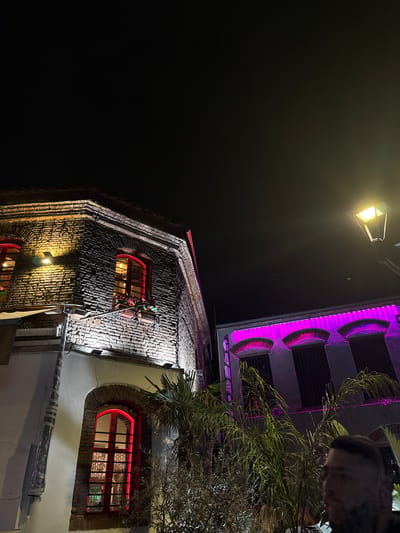 The image, taken at night, shows a street scene in Thessaloniki, Greece. The perspective is tilted, focusing on two buildings. The lower building has brickwork and windows with red accents. One window is decorated with what appears to be a Christmas garland. A streetlight illuminates part of the building. The upper building is lighter in color and illuminated with purple lights highlighting its architectural details. A person's head is partially visible in the upper right corner, and there are leafy trees and branches in the same area. The sky is dark and clear.