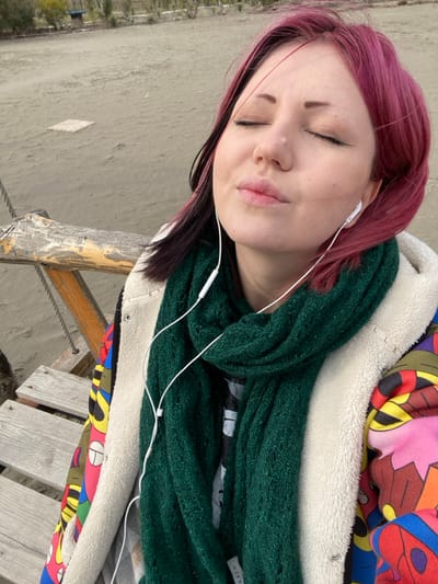 A single individual, appearing female, is depicted in a medium shot on a sandy beach in Ulcinj, Montenegro. The subject's head is tilted slightly upwards, eyes are closed, and a relaxed expression is present on her face with lips gently pressed together. Her hair is shoulder-length, primarily magenta-pink with darker sections underneath or at the roots. White wired in-ear headphones are inserted into both ears, with the wires visible descending over her chest. She wears a dark green knitted scarf wrapped around her neck, a dark garment visible beneath the scarf, and an outer jacket. The jacket features a cream-colored faux fur collar and lining, with a visible interior section displaying a bright, abstract pattern in red, yellow, blue, and black. To the immediate left, a weathered wooden structure, consistent with a bench or swing frame, is partially visible with horizontal planks and a rounded support beam. The background consists of a broad expanse of light brown sand, exhibiting subtle undulations. A small, light-colored square object is partially embedded in the sand in the mid-ground. Further in the distance, a sparse line of vegetation and an indistinct structure are visible under diffused lighting.