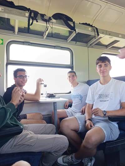 Four young men are captured inside a train carriage during a bright day, likely in Stambolovo, Bulgaria. The scene suggests a relaxed journey or social gathering.
On the left side of the frame, a man in a green hoodie and light beige pants is partially visible, with only his torso and legs seen. Next to him, a young man wearing glasses and a black t-shirt smiles broadly towards the camera. He sits across a small table, which holds a clear water bottle and a black, round object, possibly a portable speaker.
Across the aisle, two other young men are seated. The one closer to the table, also smiling, wears a white t-shirt with a faded blue graphic that includes the text "St. 1992" and other indistinct lettering. The man on the far right, with a more direct gaze at the camera, wears a white t-shirt featuring a black graphic with the prominent text "HUGO" and "CREATIVE DEPARTMENT". He also wears a silver watch on his left wrist. The men are dressed casually in t-shirts, shorts, and sneakers, suitable for warm weather travel.
Bright sunlight streams through the large train windows, illuminating the interior. Overhead luggage racks store black backpacks; one strap clearly displays the brand "VIVESS". Numbers "28", "23", and "17" are visible on different sections of the overhead compartments, likely indicating seat or storage area designations. The overall atmosphere appears cheerful and relaxed, with the men enjoying their time together on the train.
