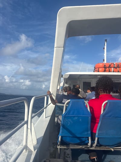 A view from the upper deck of a white passenger vessel shows multiple individuals seated, traveling across open water. The vessel is in motion, generating a white wake along its port side, with choppy dark blue water visible. A distant landmass, appearing as a dark green island or coastline, is visible on the port horizon under a partly cloudy blue sky.
In the immediate foreground, the back of a person with dark, curly hair wearing a red shirt is visible, seated in a blue canvas-backed chair. To their left and slightly forward, another individual wears a dark blue shirt with the partial text "GOVE" visible on the back, and a dark head covering. This individual's right arm is bent at the elbow, hand gripping a white railing, revealing a silver-toned wristband or watch. Further forward, other passengers are seated in similar blue chairs, their backs or profiles visible.
The vessel itself features white railings along the deck and a white structural arch or frame dominating the upper right portion of the image. A "NO SMOKING" sign is affixed to a white cabin wall, and another sign partially reads "UNTIL VESSEL DOCKED." At the stern, stacked orange life rafts/buoys are visible, along with a white mast topped by red navigational lights. The scene suggests travel by sea, consistent with inter-island transport in the British Virgin Islands, such as routes involving Spanish Town.