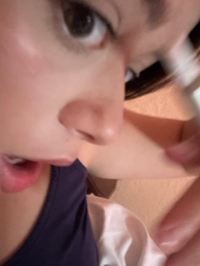 The image is a close-up, low-angle, indoor, candid shot of a person's face. The person's left eye, nose, and mouth are visible, with their mouth partially open and their lips glossy. They are wearing a dark-colored garment. The background consists of soft, blurred tones of peach and pink, suggesting an indoor setting, possibly a bedroom or living area. There is no discernible connection to Garmen, Bulgaria.