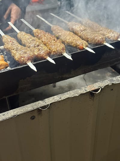 Six skewers of ground meat, possibly kebabs, are actively grilling over a charcoal fire, emitting visible white smoke. The meat on the skewers appears to be minced or ground, with some browning evident on the surfaces. A seventh, smaller, irregular food item, lighter in color, is visible on the far left of the grill. The skewers rest on a dark, heavy-gauge metal grill structure, likely a mangal. A person's hand, partially visible in the upper left, wearing a dark sleeve, is present near the cooking area. Below the main grill structure, a section of a light-colored, possibly corrugated metal or concrete barrier is visible, showing signs of wear and a single screw attachment. The scene is located in the city of Pap, Uzbekistan.