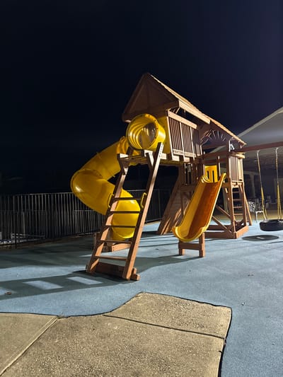 A playground structure dominates the scene at night. It features a wooden frame with a small house-like section on top. Yellow tube slides and a traditional open slide are attached to the structure. A wooden ladder provides access. The playground sits on a blue, soft surface, bordered by a concrete edge. The background is dark, with minimal illumination highlighting the playground equipment.