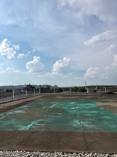 The media file displays a tilted view of a rooftop or elevated walkway, with a clear blue sky and scattered white clouds dominating the left side of the frame. The right side of the image features a concrete surface with distinct patches of weathered teal-colored paint, suggesting it might be a sports court or a decorative element. A narrow strip of small, light-colored pebbles lines the edge of the concrete.

In the background, a cityscape unfolds with buildings and trees, partially obscured by a distant mountain range. Various structures, including what appear to be solar panels, are visible along with fences and utility poles. The overall scene suggests an outdoor urban or semi-urban environment.

The lighting indicates it is daytime, likely late morning or early afternoon, given the bright sunlight and the clarity of the sky. The weather appears to be fair and pleasant. No people are visible in the frame. The image does not contain any visible text. The perspective is from a high vantage point, looking down and outwards.