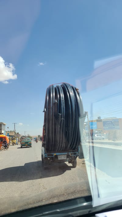 The image, when viewed upright, depicts a bustling street scene in Kuchlak, Pakistan, on a sunny day. The main subject is a light blue truck overloaded with a substantial quantity of coiled black pipes, likely used for water or gas infrastructure. These pipes are stacked high and wide on the truck bed, secured with visible straps.

The truck is positioned on a paved road, traveling away from the viewer. Its rear license plate is clearly visible, bearing the text "WAD 768" with "PAKISTAN" written underneath, indicating its registration. The bright sunlight casts distinct shadows on the road, suggesting it is daytime, possibly around noon. The sky is a clear, bright blue with a few scattered white clouds.

Along the right side of the road, a row of commercial buildings lines the street. Several billboards with text in Urdu are displayed on these structures. One prominent sign reads "رتیں" (Raten) and "پراپرٹی کی دیوار" (Property ki Deewar), while another below it states "صبا برادر" (Saba Brothers), indicating local businesses, possibly related to real estate or construction.

Further down the street, various other vehicles are present, including a green minivan or pickup truck, several auto-rickshaws, and motorcycles, some carrying passengers. The scene portrays typical daily traffic and commercial activity in a South Asian urban or semi-urban environment. The photograph appears to have been taken from the interior of another moving vehicle, with reflections visible on the glass surface through which the shot was composed.