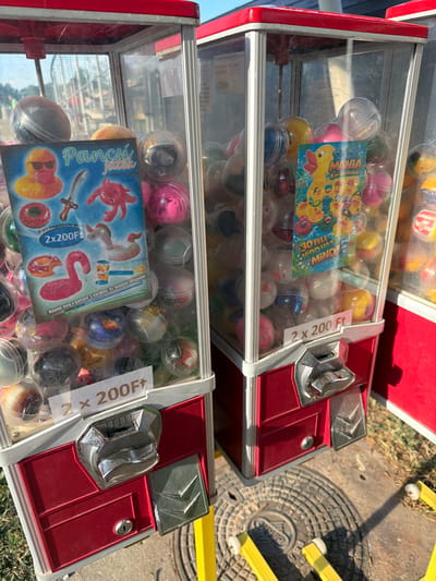 This media file shows two red and silver vending machines filled with colorful plastic capsules. Each capsule appears to contain a toy. One machine has a label featuring cartoon ducks and the text "DUCK MANIA" and "KACSA MANIA". The other machine has a label with cartoon sea creatures and the text "Pancsi jatek" and "Felfújható vizi jate". Both machines have a price displayed as "2 x 200Ft". The machines are positioned outdoors on a paved surface, with grass visible in the background. A manhole cover and yellow metal supports for some unseen structure are also present. The lighting suggests it is daytime, possibly sunny. The overall scene depicts a street or public area where these vending machines are located, likely for children's toys.