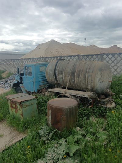 The image depicts a derelict blue truck with a large, rusty, cylindrical tank trailer attached, located in Яльгелево, Russia. The scene appears to be a storage or abandoned area, overgrown with tall grass and weeds. To the left of the truck, there is a rusty green metal box and a cylindrical rusty barrel. Behind the truck is a tall mound of sand and a chain-link fence. A cell tower is visible on top of the sand. The sky is overcast with heavy clouds.