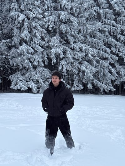 In Le Hohwald, France, a male individual stands in a snow-covered outdoor environment. The subject is positioned centrally, facing forward but looking slightly to his left. He has dark hair and a light mustache. He is dressed in a black puffer jacket, with his hands in the pockets, and dark trousers. The snow covers the ground to approximately mid-calf level on his legs, with snow visible adhering to the lower portions of his trousers. In the background, a dense forest of evergreen trees is visible, with branches heavily coated in white snow or hoarfrost. The sky is overcast.