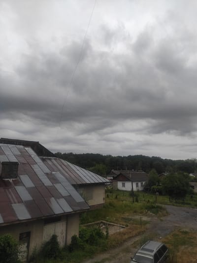 A gray, overcast sky looms over a rural scene in Kostopil, Ukraine. In the foreground, the corrugated metal roof of a building is visible, with a brick chimney on its left. To the right, a narrow, dark line stretches across the sky. Further in the background, several small houses with pitched roofs are scattered among trees and overgrown vegetation. A dirt road winds through the scene, with a dark-colored station wagon parked on it. The overall impression is one of a quiet, possibly somber, day in a village.