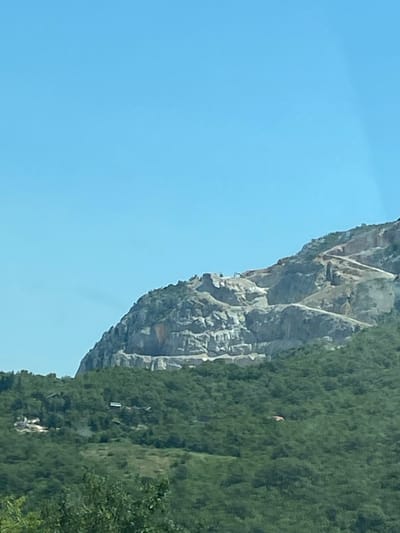 An aerial view shows a quarry carved into a steep, rocky mountainside in Mišići, Montenegro. The quarry exhibits numerous terraces and excavation sites, indicating ongoing or recent activity. The surrounding lower slopes are covered in dense green foliage. Several small, dark-colored buildings are visible scattered within the trees on the lower slopes. The sky above is a clear, bright blue.