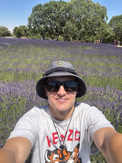 A man in a light gray t-shirt and a dark gray bucket hat takes a selfie in a lavender field. He is wearing dark sunglasses and has braces on his teeth. The t-shirt has a red graphic of the number "2023" with an image of a tiger next to it. The background is filled with rows of purple lavender plants under a bright blue sky. To the left of the lavender field, there are large green trees. The time of day appears to be sunny and warm, suggesting it is daytime. The overall impression is a cheerful and relaxed moment captured during an outdoor excursion. The setting is a lavender farm in the Golan Regional Council, Israel.