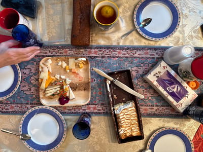 The image presents an overhead view of a dining table in Binyamina, Israel, set for a casual gathering. The table is draped with a pale yellow patterned tablecloth, overlaid by a central fabric runner featuring blue, pink, and red patterns.

On the left side of the frame, a pair of human hands with painted fingernails reaches towards a blue patterned glass, positioned near an empty white plate with a blue decorative rim and a gold edge. Above this area, a red mug containing a dark liquid rests beside a black rectangular object. An empty clear plastic container lid is situated further right.

Centrally, a rectangular wooden serving board displays a variety of partially consumed cheese pieces, along with small red and green edible garnishes. Above this board, a brown rectangular loaf of bread is visible. To the right of the bread, a white and blue mug holds a liquid and a lemon slice. An empty white plate with a blue rim and a silver spoon rests above the table runner.

On the right side of the runner, a dark rectangular dish contains a dessert with a golden, wavy-patterned meringue topping, accompanied by a white-handled knife. Below this, a floral-patterned box of "Soft Tissue" is visible, featuring a purple label. Adjacent to the tissue box are a stack of clear plastic disposable cups, a white container with a floral design, a red lidded container, and an orange bottle cap. An additional blue patterned glass and another empty white plate with a blue rim and a silver spoon are visible in the lower left portion of the image. A black patterned cup is partially visible in the lower right.