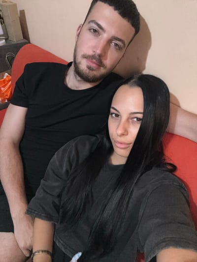 A couple is sitting on a red sofa in what appears to be a living room. The man on the left has short, dark hair and a beard, wearing a black t-shirt. The woman on the right has long, dark hair and is wearing a dark grey t-shirt. They are both looking towards the camera, with the man's arm around the woman. The background is a plain, light-colored wall, with some shadows cast on it. The overall impression is of a casual, intimate moment captured in a domestic setting. There is no specific indication of the location in Каменар, Bulgaria, but the style of the room suggests it is a residential interior.