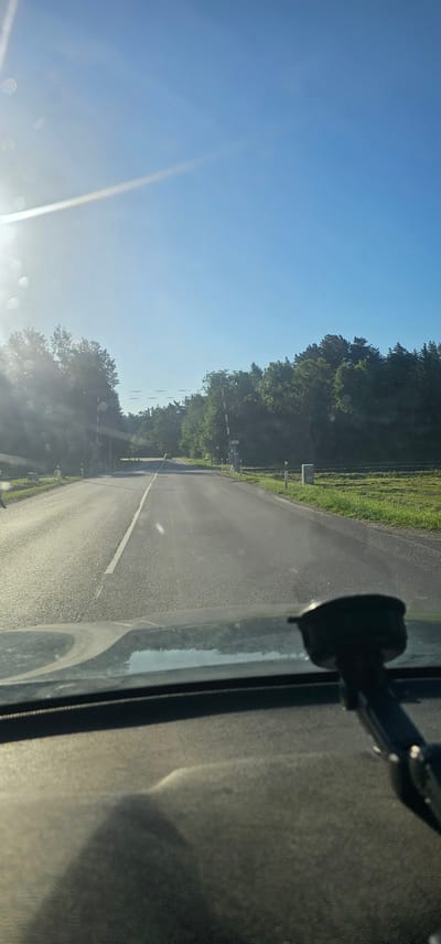 The image shows a view from inside a vehicle looking out through the windshield at a paved road. The road is lined with trees on both sides. There are two railway crossing barriers visible, one on each side of the road. The sky is blue and clear with the sun shining brightly, causing lens flare and reflections on the windshield. The overall environment appears to be rural, with green fields and dense trees. The dashboard of the car is visible in the foreground, with a dark object, possibly a phone mount, attached to it.