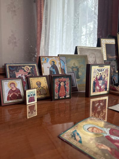 The image depicts a collection of small religious icons displayed on a polished wooden surface, likely a table or shelf. The icons feature traditional religious imagery, including depictions of Jesus Christ, Mary, and various saints. Some icons are framed and stand upright, while others are smaller and lie flat. The background shows a blurred reflection, possibly of a window with curtains, suggesting an indoor setting. The lighting is soft and diffused. There are no people visible in the image. The overall scene suggests a personal collection of religious artifacts. The user's request for location in Tutaev, Russia, cannot be verified from the visual information provided in the image.