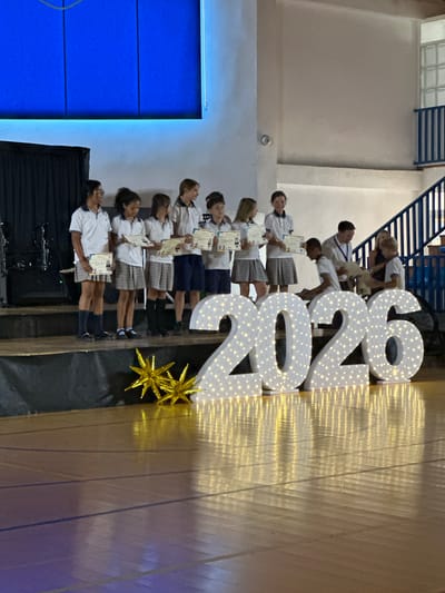 In San Pedro De Alcántara, Spain, an awards ceremony is taking place on a stage. A group of children, dressed in school uniforms, stand on a raised platform, each holding a certificate. Behind them, two adults are presenting certificates to other children. In front of the stage, large illuminated numbers spell out "2026." The floor is a polished wooden surface, reflecting the lights. The backdrop includes a large blue screen with some lighting effects and black curtains. There are also golden star decorations on the stage.