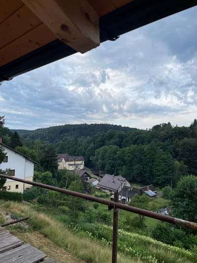 This image depicts a view from a raised wooden structure, looking out over a semi-rural landscape. In the foreground, the edge of the wooden structure is visible, with planks of wood. A rusty metal railing runs horizontally across the mid-ground, supported by vertical metal poles. Beyond the railing, a grassy slope descends, dotted with white wildflowers and taller grasses.

Further into the scene, several buildings are visible. A large, white building with a light yellow trim is situated on the left side of the frame. Several smaller residential buildings with pitched roofs, some appearing to be made of brick and others of wood, are clustered in the middle and right portions of the image. One building has a distinctive orange chimney. A dark-colored vehicle, possibly a car or van, is parked in a driveway or yard near a few of the houses.

The background is dominated by a dense, verdant forest that stretches across rolling hills. The trees are lush and green, indicating a temperate climate. The sky above is overcast with scattered clouds, suggesting a partly cloudy day. The overall impression is one of a tranquil, wooded area, likely a village or small town on the outskirts of a more populated area. The location is identified as Lhotka, Czechia.