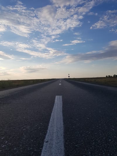The image shows an empty asphalt road stretching into the distance under a partly cloudy sky. The road has a white dashed line down the center and a solid white line on the right edge. The landscape on either side of the road is flat and covered with dry grass and some scattered trees in the far distance. A single signpost with a circular element is visible on the right side of the road in the middle distance. The lighting suggests it is either early morning or late afternoon. The scene appears to be in a rural or steppe environment. Without further context or visible landmarks, the specific location within Дөлен ауылдық округі, Kazakhstan, cannot be determined solely from the visual information in the image.