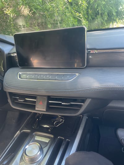 The image shows the interior of a car, likely a modern SUV, with a large central infotainment screen. Below the screen are a series of touch-sensitive buttons for climate control, including icons for A/C, air circulation, defrost, and fan speed. A red hazard light button is prominently placed between two air vents. The gear shifter is a rotary dial with illuminated indicators for Park (P), Reverse (R), Neutral (N), and Drive (D). A set of car keys are hanging from a hook near the shifter. The dashboard is textured with a patterned material and features blue stitching along a seam. The exterior environment, visible through the windshield, consists of green foliage and dappled sunlight, suggesting an outdoor, possibly daytime setting. The image was taken from the driver's perspective.