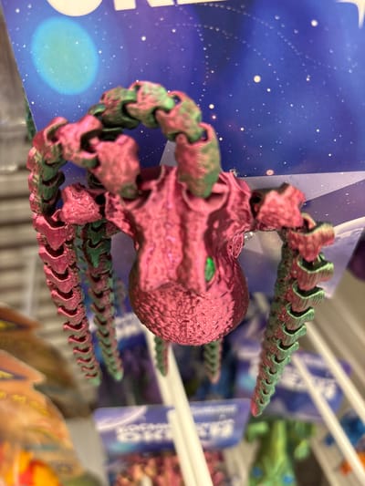 This media file showcases a close-up, top-down shot of a pink and green articulated toy, likely a 3D printed octopus or similar creature, resting on a surface. The toy's segmented body and tentacles are detailed, with a textured, almost metallic sheen. One of the creature's eyes is visible, a bright green gem-like feature.

The background is a blurred display of what appears to be merchandise. Behind the toy, a blue card with white speckles, resembling a starry sky, is visible. To the right and further back, other items are displayed on shelves, some in purple and blue packaging with white text. Some blurred green and orange objects are also visible in the background, suggesting a retail environment.

The lighting appears to be artificial and diffused, casting soft shadows. No people are visible in the frame. The setting suggests an indoor retail space, possibly a toy store or a novelty shop, given the display of various items. The focus is entirely on the toy, highlighting its intricate design and dual-color construction. The image's shallow depth of field blurs the background, emphasizing the toy as the primary subject. There is no discernible time of day or weather information. No readable text is visible on the toy or the immediate background elements.