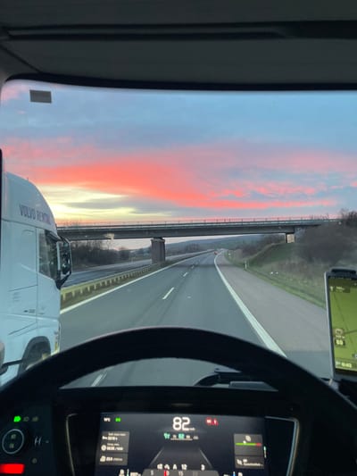 A forensic examination of the image reveals a multi-lane highway scene at dawn or dusk in the city of Wallhausen, Germany, observed from the driver's perspective inside a vehicle.

The sky exhibits vivid pink and red stratiform clouds transitioning to light blue towards the zenith. In the foreground, the upper portion of a steering wheel is visible, below which a digital dashboard display shows a speed of 82 km/h, an outside temperature of 1°C, a time of 02:18, trip mileage of 22.4 km, fuel consumption of 22.4 l/100km, and a projected remaining distance of 897 km. Multiple green indicator lights are active on the dashboard's left side, alongside a single red indicator light at the bottom left. A navigation device mounted on the dashboard's right side displays a map, the number "80" (likely a speed limit), and "0:42" (possibly a remaining time).

To the immediate left, a white Volvo 500 series truck with "VOLVO RENTAL" branding is visible in an adjacent lane, moving parallel to the viewing vehicle. The highway itself consists of at least two lanes for the direction of travel, with clear white lane markings and a concrete barrier separating it from the opposite direction of traffic. A grey concrete overpass bridge, supported by two visible rectangular pillars, spans the highway approximately 200 meters ahead. Beyond the bridge, the highway curves gently to the right, with sparse, leafless vegetation on rolling hillsides. Distant vehicle lights are faintly visible on the highway ahead. No individuals are directly visible within the frame.