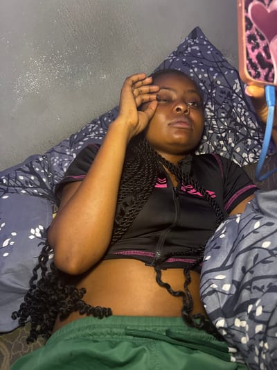A young adult female is depicted lying on her back indoors in Gbongan, Nigeria. She has dark skin and long, black braided hair with curly ends. She is wearing a black short-sleeved crop top with a front zipper and pink trim, exposing her midriff, and green athletic shorts. Her right hand is raised, with fingers positioned near her right eye, while her left hand holds a mobile phone. The phone case is pink with a black leopard print and pink heart embellishments, and a blue charging cable is plugged into its bottom. She appears to be looking towards the phone with her eyes partially closed. She is resting on a bed covered with dark blue bedding featuring a white tree branch and leaf pattern. The wall behind her is dark gray with subtle white speckles.