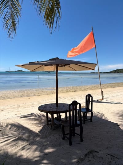 The image depicts a tranquil beach scene in Baan Thong Krut, Thailand. In the foreground, a round wooden table with two dark wooden chairs sits beneath a large, light-colored umbrella, casting shadows on the sand. The umbrella is positioned to provide shade over the seating area. To the right of the umbrella, a bamboo pole stands vertically, supporting a bright orange flag that flutters in the breeze.
The sandy beach gently slopes down to the calm, clear blue water. In the distance, the sea is dotted with a few small boats, and a lush green island with a distinctive peak is visible on the horizon. The sky is a brilliant, cloudless blue, characteristic of a sunny day. Palm fronds hang from the top left of the frame, adding to the tropical ambiance. The overall atmosphere is one of peaceful seclusion and natural beauty.