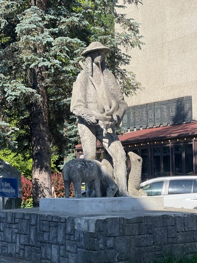 The image shows a concrete sculpture of a shepherd with a dog and sheep, located in front of a building with a dark metal facade and a red roof. The sculpture depicts a man in a hat, holding a staff and a musical instrument, standing next to a sheep and a dog. The man's clothing appears to be made of rough, textured material, and his facial features are indistinct. The sheep and dog are also rendered in a stylized, simplified manner. Behind the sculpture, a large tree with blue-green foliage is visible, as is a section of the building's exterior wall. A white car is parked to the right of the sculpture. A blue signpost with Polish text that reads "Skwer Andrzeja Winiarczyka Galicy" is positioned to the left of the sculpture. The sculpture is placed on a stone pedestal, which is supported by a low, segmented stone wall. The overall scene suggests an outdoor public space, likely a park or square, in a city. The sculpture is a public art piece commemorating a local figure or aspect of regional culture.