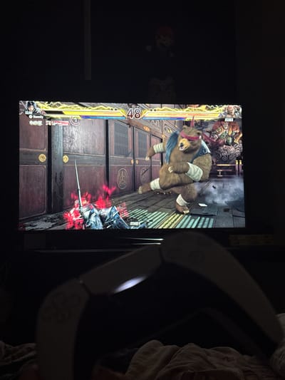 The image shows a television screen displaying a fighting game, possibly Tekken 7, where Kuma the bear is fighting another character. The game is set on a stage with a Japanese-style building as a backdrop. The health bars for both characters are visible, with numerical values. Adjacent to the TV is what looks like the headband portion of a VR headset. The environment appears to be a dimly lit room, perhaps a living room or bedroom. There is bedding material in the top right corner. The image does not provide any information about the location of Bergen, Norway.