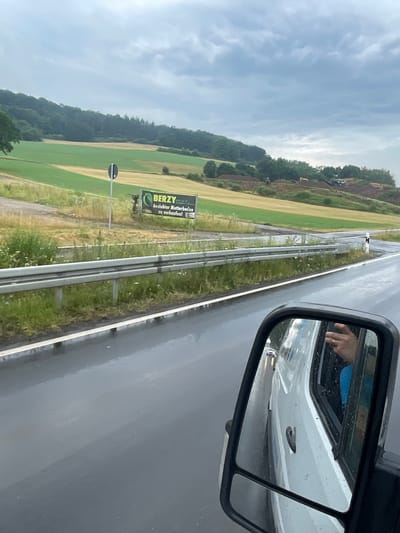 The image is a forward-facing exterior shot of a white van's side mirror, taken from inside the van. The mirror reflects the van's exterior, the road, and the surrounding landscape under an overcast sky. In the distance, there are rolling hills with agricultural fields in shades of green and yellow, and a forested area. A road runs along the edge of the fields, with a metal guardrail. A billboard advertising "BERZY" and "Gesiebter Mutterboden zu verkaufen!" (Sifted topsoil for sale!) is visible on the side of the road. The road surface is wet, indicating rain. In the mirror's reflection, the hand of the van's driver, wearing a ring, is visible, along with a blue shirt. Further down the road, a white car is seen through the van's window. The scene appears to be a rural area outside Wartenberg, Germany.