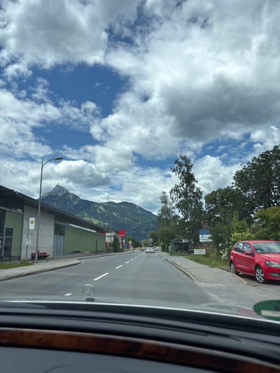 The image is a street-level view of a road in Reutte, Austria, on a cloudy day. The perspective is from inside a car, looking forward. On the left, there is a long, modern building with a grey facade and green panels, partially obscured by trees. A bus stop sign with a 'H' symbol is visible on the sidewalk in front of this building. A red bench is also present on the sidewalk. Further down the road, a large mountain range dominates the background, with trees and vegetation on its slopes.
The road itself is asphalt with white lane markings. Several cars are driving on the road, including a red Volkswagen Polo parked on the right side of the street. Other vehicles, including a white van, are seen driving in the distance. There are also streetlights and signs along the road. On the right side of the road, there is a grassy verge with trees and some small structures that appear to be shops or stalls. Signs for "Spar Markt" and "Sport Center" are visible.
The sky is partly cloudy, with large white and grey clouds against a blue sky. The overall impression is a typical European town scene with a backdrop of dramatic natural scenery.