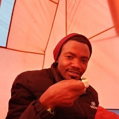 Gilman's Point, Tanzania: A single adult male is depicted in a medium shot, centered within an orange-toned interior, likely a tent or shelter. He possesses dark skin, short dark hair, and a trimmed beard, wearing a dark red knit beanie and a black quilted jacket with a visible 'BB' logo on the left chest. His right hand holds a small piece of white food, possibly a boiled egg or fruit, garnished with green elements, positioned near his mouth. He is looking directly at the camera with a slight smile. The background shows orange fabric walls with structural lines, and a light blue section is visible in the upper left quadrant, indicating an external view or window.
