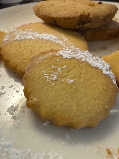 A close-up view displays a collection of light brown cookies presented on a white, possibly ceramic, surface. The foreground features a round, scalloped-edge cookie heavily dusted with white powdered sugar, which also scatters across the white surface. Behind it, another similar cookie is partially visible, also exhibiting powdered sugar. Further back, two more cookies are stacked; the upper one is round with a smooth edge and appears to contain dark inclusions, possibly chocolate chips, while the lower one is largely obscured. To the right, a fragmented portion of an additional cookie or crumbs is visible. The scene is located in Andratx, Spain. No individuals or active interactions are depicted.