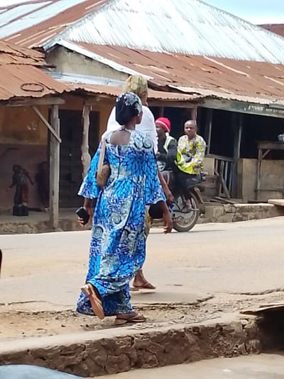 This media file depicts a street scene in Iseyin, Nigeria, under an overcast sky. The primary subjects are several people, including a woman in the foreground wearing a vibrant blue and white patterned dress and a headscarf. She is walking on a paved road next to a raised concrete curb. She holds a dark-colored mobile phone in her left hand and has a tan purse slung over her right shoulder.

Behind her, another person, partially visible, walks alongside. Further in the background, two individuals are on a motorcycle. One person is driving, wearing a yellow and green patterned shirt, and the other is a passenger wearing a red headscarf and a dark jacket with a reflective vest over it.

The setting includes a row of buildings with corrugated metal roofs, some showing signs of rust and age. The structures appear to be shops or homes, with wooden supports and exposed walls. In the far left background, a figure is visible in what might be an open doorway, standing on elevated platforms.

The activity appears to be a normal day with people moving along the street, suggesting ordinary daily life rather than a specific event. The overall atmosphere is somewhat muted due to the cloudy weather. There is no visible text in the image. The scene is indicative of a town or market area in Nigeria, with the details of the buildings and the clothing of the people providing location cues.
