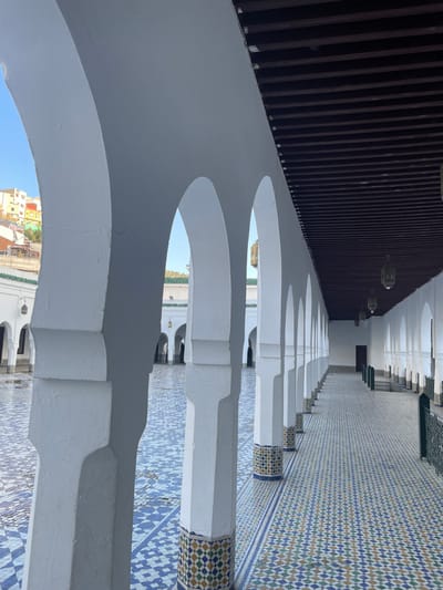 An architectural scene is depicted within a large complex in the city of Moulay Idriss Zerhoun, Morocco, likely the Mausoleum of Moulay Idriss I. The image captures a covered walkway (riwaq) with a central courtyard to its left.
The foreground is dominated by a series of white, thick, horseshoe-shaped arches forming the right side of the walkway. The walkway extends into the background on the right, defined by a receding line of identical arches and a low dark railing. The ceiling of the walkway consists of dark brown horizontal wooden beams, from which several ornate, dark metal lanterns are suspended at regular intervals. The floor of this walkway is paved with intricate mosaic tiles (zellij) featuring a repeating pattern of blue, green, brown, and white diamonds. The base of each pillar supporting the arches is adorned with a band of similar mosaic tilework.
To the left of the arches, an open, expansive courtyard is visible, paved with light blue and white geometric mosaic tiles, appearing slightly damp or reflective. Beyond the courtyard, other white structures with green-tiled roofs and arched doorways are visible, forming part of the surrounding complex. In the far background, above the complex and through the arches, a steep hillside densely covered with multi-colored buildings (yellow, orange, white, and red) is visible under a clear blue sky, indicating the surrounding city landscape. No individuals are present in the scene.