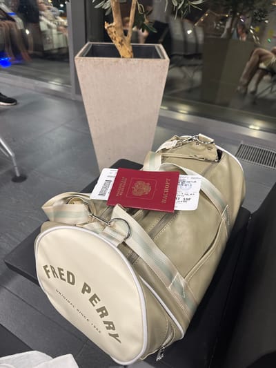 A light-colored duffel bag with "FRED PERRY ORIGINAL SINCE 1952" printed on it sits on a dark surface. A red Russian passport and a boarding pass are placed on top of the bag. In the background, a large concrete planter contains a tree. The scene appears to be in an airport or a similar transit area, given the presence of a boarding pass and a passport, and the reflections in the glass windows suggest a public waiting area.