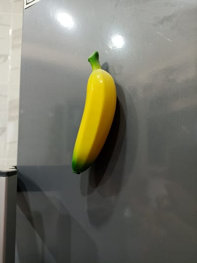 A glossy, banana-shaped refrigerator magnet is affixed to a dark grey, metallic surface, consistent with a refrigerator door. The magnet features a bright yellow body with green coloration at its stem and the opposite end. Reflections from light sources are visible on both the magnet and the metallic surface, which exhibits minor surface abrasions. To the immediate left, a portion of a lighter-colored, possibly stainless steel appliance or counter edge is visible. In the background on the far left, a wall surfaced with light-colored, square tiles is partially discernible. No individuals or discernible actions are depicted. The specific location within Mu'adammiyat ash-Sham, Syria, cannot be determined from the visual evidence provided.