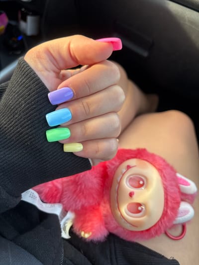 A person's hand, adorned with pastel-colored acrylic nails in shades of lavender, sky blue, mint green, and pale yellow, is held up in the foreground. The nails are square-shaped and appear to be newly manicured. The person is wearing a black long-sleeved garment. In the background, partially visible, is a fluffy pink toy with a doll-like face, featuring large pink eyes and a small pink heart-shaped nose. The toy has bunny-like ears. The setting appears to be inside a vehicle, indicated by the dark interior and what looks like a car dashboard or similar surface in the upper right corner. The lighting suggests it is daytime, possibly sunny. There is no indication of the location being in Λούρος, Greece, or any specific city.