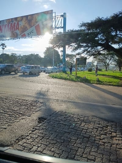 The image presents an outdoor scene at a multi-lane urban intersection or roadway in Harare, Zimbabwe. The time of day appears to be mid-morning or late afternoon, indicated by direct sunlight causing significant lens flare and strong shadows.

In the immediate foreground, the asphalt road surface is severely degraded, featuring extensive cracking, an area of exposed, irregular paving stones or broken tarmac, and a visible pothole. A dark vehicle, possibly a pickup truck, is partially visible in the lower left, followed by a white van and multiple other light-colored passenger vehicles queued in traffic, suggesting congestion on the left-hand drive road.

Dominating the left-center is a large, tall billboard supported by a blue metal frame, displaying a KFC advertisement featuring fried chicken and the partial word "NYAMA." The sun is positioned behind this billboard, creating a bright glare.

To the right of the roadway, a wide, grassy median strip or verge contains several mature trees with dense foliage, one particularly large tree extending its branches across the upper right quadrant. At least three individuals are observed standing on this grassy area; two are near a dark rectangular signpost, and a third is further right, partially obscured by a tree. Their postures suggest waiting or observation. The background includes further trees, suggesting a green urban environment.