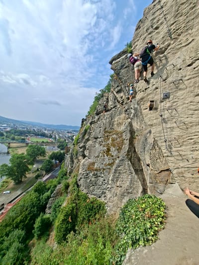 In Děčín, Czechia, a rock climbing activity is taking place on a sheer rock face overlooking a cityscape. Three individuals are visible on the rock. The uppermost climber is a male wearing a black t-shirt, blue shorts, and an orange helmet. He is assisted by a second climber, a female in a purple top and light-colored shorts, also wearing a helmet. Below them, a third, smaller individual, likely a child, is ascending the rock face, wearing a blue and white outfit and a helmet. Metal rungs are embedded into the rock face, serving as handholds and footholds for the climbers, with safety ropes and carabiners visible.

The rock face is a natural geological formation, with textured surfaces and patches of moss and vegetation. In the background, a panoramic view of Děčín unfolds. A river, likely the Elbe, flows to the left, spanned by a bridge. The cityscape features buildings, railway tracks, and roads. Lush green trees and vegetation surround the base of the rock formation and extend into the city, creating a contrast between the natural and urban environments. The sky above is partly cloudy, with patches of blue sky and white clouds. The overall scene captures an adventurous outdoor activity set against a scenic urban and natural landscape.
