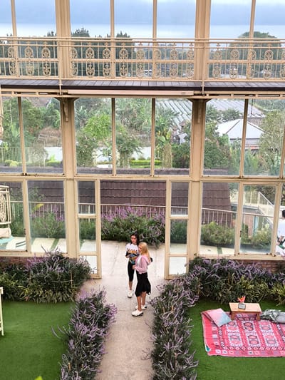 The image captures an elevated view of a garden area within a building or greenhouse-like structure, presumably located in Candikuning, Indonesia, given the presence of tropical foliage in the landscape visible through the windows.

Two young women stand in the center of a paved pathway lined with lavender bushes. One woman has dark hair and wears a white t-shirt, black shorts, and white sneakers; the other has blonde hair and wears a pink sweater, a black skirt, and white sneakers.

The path is flanked by artificial turf on either side, creating small garden sections. To the right of the path is a picnic setup on the turf. This includes a red patterned rug, several pillows, and a small wooden table with a vase of flowers.

The entire scene is framed by the structure of the building, which consists of large windows and ornate metal railings, giving a view of the surrounding lush landscape, including a tiered landscape with homes, trees, and cultivated fields. The upper portion of the structure has a balcony with ornate metal railings. The lighting appears to be overcast or diffused, which creates an even distribution of light throughout the scene.