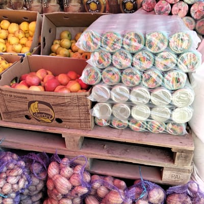 The image depicts a street market scene in Cupcini, Moldova, with a focus on various food items displayed for sale. The foreground features sacks of potatoes, some in purple netting and others in lighter colored netting. Behind the potatoes, stacked wooden pallets support several cardboard boxes. One box, labeled "PRODUCT OF ECUADOR," contains red and yellow apples. Another box, also containing apples, is visible to the left.
Further back, a stack of individually wrapped items, possibly cups or lids, is covered in plastic. The wrappers have colorful designs and some Cyrillic text, suggesting they are intended for the local market. In the background, bags of coffee or similar products are visible, also with Cyrillic lettering.
The scene suggests a daytime market, likely during business hours. The overall impression is one of commerce and fresh produce available to consumers. There are no people visible in the image, focusing entirely on the goods for sale and their arrangement. The presence of produce from Ecuador alongside local items indicates trade and a diverse offering at the market. The visible text includes brand names like "derby" and "EPAL" on the packaging and pallets, as well as product information in Russian.