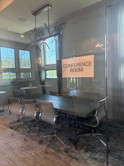 This forensic image depicts the interior of an unoccupied modern conference room, located in Orlando, United States, viewed through a reflective clear glass partition. A prominent rectangular sign on the glass states "CONFERENCE ROOM" in black text, with supplementary smaller text and braille underneath. The room contains a large, dark-finished rectangular conference table. Eight contemporary office chairs with light-colored seating surfaces and chrome five-star bases on casters are arranged around the table; six are clearly visible. Overhead, two distinct chandeliers illuminate the space: a linear, abstract metal fixture positioned directly above the conference table, and a larger, rectangular cage-style chandelier located further into the room. Recessed ceiling lights and a circular ceiling speaker are also present. The left wall is dominated by multiple large windows, offering a partial exterior view of green trees under natural daylight. Light-colored curtains are drawn along the right side of the room. The flooring is a dark, patterned carpet. Beyond the main conference area to the left, two small, round white pedestal tables, each adorned with a white floral arrangement, are visible. No individuals are present, and no actions are occurring within the frame.