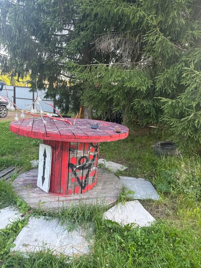 In the city of Lyady, Russia, an outdoor scene features a repurposed red cable spool table with graffiti. The table is adorned with a string of bare light bulbs hanging from branches, with some bulbs illuminated. A black smartphone rests on the table's surface. A white sign with black arrows pointing in opposite directions is placed beside the table. The area around the table is covered with grass and interspersed with large, irregular concrete slabs. In the background, a large evergreen tree dominates the landscape, with a section of a metal fence and a vehicle visible to the left. To the right, a discarded tire lies in the overgrown grass.