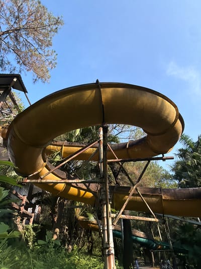 An outdoor image taken in Thủy Xuân Ward, Vietnam shows a yellow, cylindrical water slide that loops vertically with visible rivets. The slide is supported by an aging, rusted metal framework that consists of vertical and diagonal supports. The sky is visible in the background, along with trees and foliage. Parts of a dilapidated roof are visible at the bottom. Vegetation is present on the right side of the image.