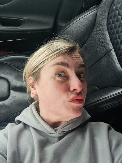An adult female is depicted from the chest up, positioned within the passenger compartment of a vehicle. She has blonde hair, a light complexion with visible freckles, and wide, dark-rimmed eyes. Her eyebrows are raised, causing horizontal creases on her forehead, and her lips are pursed and tinted reddish, forming a "duck face" expression. A small, silver-colored stud earring is present in her left earlobe. She wears a light grey hooded sweatshirt with a visible drawstring at the neckline. The background consists of dark-colored, likely black, leather car upholstery, featuring a quilted or diamond-stitch pattern and perforated sections, consistent with a vehicle interior. A dark car door panel with a recessed handle is visible to the left. The specific geographic location within the city of Sasovo, Russia, cannot be determined from the visual information presented in the image.