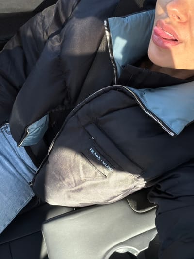 A close-up, high-angle image depicts a person seated inside a vehicle in Villeurbanne, France, under direct sunlight. The subject's upper torso and the right side of their face are visible. They are wearing a black puffer jacket with a light blue interior lining, featuring a silver-toned zipper and a visible "PRADA - MILANO" patch with a triangular logo on the left chest area. Light blue denim jeans are visible on their left thigh. The person's mouth is slightly open, with their tongue partially extended, and their lips appear glossy. A dark black strap, possibly a seatbelt or bag strap, crosses their upper chest. The immediate environment includes a dark grey vehicle interior console or armrest with a visible cup holder recess on the lower right side of the frame. Strong, direct sunlight casts bright highlights across the subject's face, jacket, and the vehicle interior.