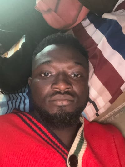 A dark-skinned adult male is depicted in a close-up, head-and-torso shot, presumably a selfie, taken in the city of Ozalla, Nigeria. He is lying down, looking directly at the viewer with a neutral expression. He possesses short, dark, curly hair with faded sides, a full beard, and a mustache. He is wearing a red knit cardigan featuring black horizontal stripes on his right shoulder/chest area and a cream, green, and red striped trim with a dark circular button on his left. The background consists of bedding or pillows: a pillow with vertical red, white, and blue stripes is visible to his right, and another with horizontal light blue and dark blue stripes is seen to his left. A dark, possibly black, fabric object is partially visible behind his head on the upper left. The lighting is low.