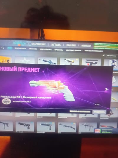 The image depicts a computer screen displaying a video game interface, specifically a loot box or item drop screen. The prominent element is a stylized revolver with a rainbow gradient, labeled as "Revolver R8 | Amber Gradient" from the "Dust 2 Collection." The background is a vibrant purple with glowing lines. Above this, the text "NEW ITEM" is displayed in large pink lettering. The top of the screen shows a menu bar with various options like "Inventory," "Loadout," "Play," "Market," and "News." The environment is that of a person viewing a computer screen, likely in a personal space, with reflections visible on the screen. There is no indication of the city of Yorkatay, Uzbekistan, or any specific geographical location.