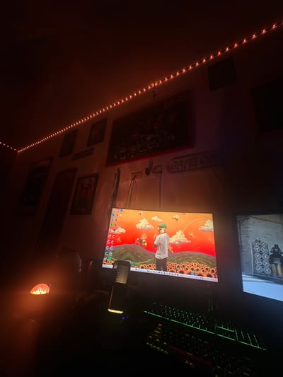 The image depicts a dimly lit room, possibly a bedroom or study, in an unknown location, as there is no indication of location within the image. There are string lights diagonally spanning the left side of the wall. The wall is decorated with several framed pictures and a "GAME DAY" sign. A computer setup is visible, consisting of two monitors, a keyboard with green backlighting, and a microphone. The main monitor displays an image featuring Tyler, the Creator. The overall environment suggests a personal space dedicated to gaming or entertainment.